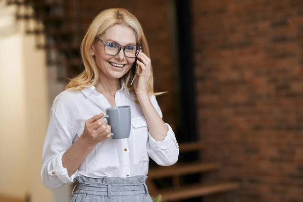 Hari yang indah. Wanita bisnis paruh baya dengan rambut pirang dan kacamata memegang secangkir kopi atau teh ketika berbicara di telepon, berdiri di rumah — Stok Foto