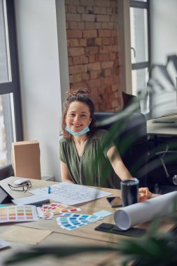 Neşeli bir kadın, koruyucu maskeli, kameraya gülümseyen bir iç mimar yeni yaratıcı proje için planlar ve renk örnekleriyle çalışırken, ofisinde masasında oturuyor.