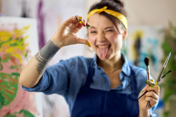 Playful female artist showing half heart love sign gesture with her hand in paint at camera while creating painting, applying colorful paints on canvas with fingers and paintbrushes