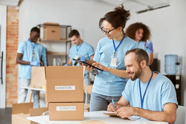 Muž a žena rádi spolupracují na projektu darování v interiéru. Tým dobrovolníků v modré uniformě si dělá poznámky, kalkuluje, třídí a balí věci v kartonových krabicích — Stock fotografie