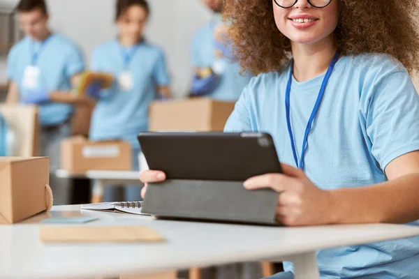 Retrato recortado de una joven voluntaria en uniforme azul usando tablet pc y sonriendo a la cámara mientras está sentada en el interior. Clasificación por equipos, artículos de embalaje en segundo plano — Foto de Stock