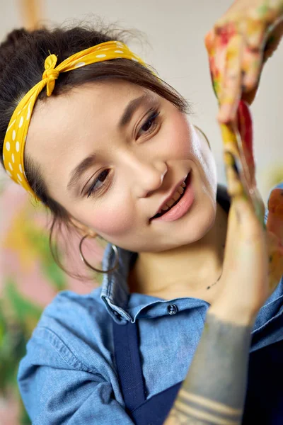 Close-up portret van vrolijke jonge vrouw, vrouwelijke schilder kijken naar haar handen in de verf, terwijl het maken van schilderen in de werkplaats — Stockfoto