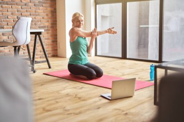 Spor giyim sektöründe aktif, olgun bir sarışın kadın. Gündüz evde yoga yaparken dizüstü bilgisayarla video dersini takip ediyor.