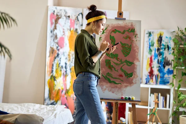 Pandangan samping dari wanita muda menggunakan smartphone-nya sambil membuat lukisan abstrak, berdiri di ruang tamu di rumah — Stok Foto