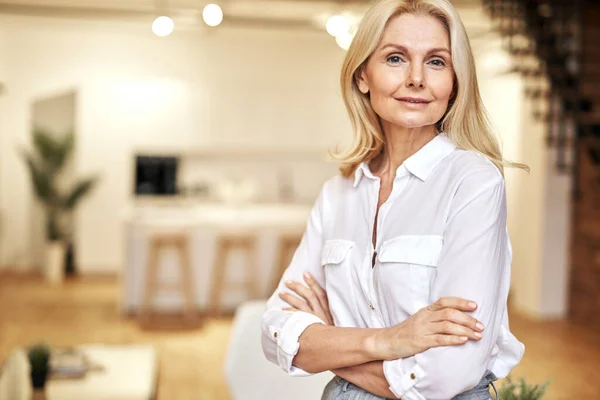 Portrait of successful mature blonde business lady looking at camera while posing, standing with arms crossed in the office — Stok Foto