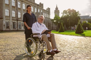 Tekerlekli sandalyedeki yaşlı engelli erkek doktorun, laboratuvar önlüğü ve gözlüklü, dışarıda ona yardım eden genç bir hemşire.