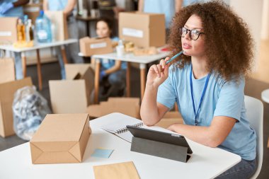 Düşünceli genç bir kadın, mavi üniformalı gönüllü, başka tarafa bakıyor, tablet bilgisayar kullanıyor ve bağış projesi üzerinde çalışıyor. Takım sıralama, arka planda paket ögeleri