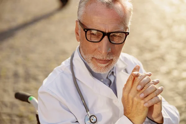 Retrato de cerca del medico anciano discapacitado en silla de ruedas usando bata de laboratorio y gafas posando al aire libre en un día soleado —  Fotos de Stock