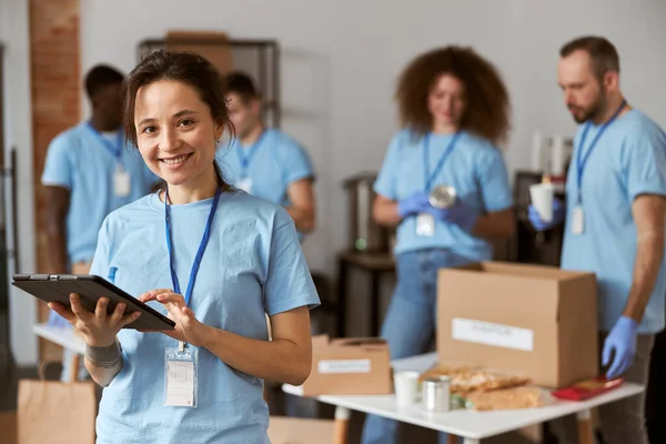 Retrato de una joven voluntaria feliz en uniforme azul usando tablet pc y sonriendo a la cámara mientras está de pie en el interior. Clasificación por equipos, artículos de embalaje en segundo plano — Foto de Stock