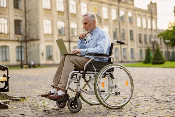 Foto lengkap pria dewasa yang sibuk, memulihkan pasien di kursi roda yang bekerja pada laptop di taman dekat klinik — Stok Foto