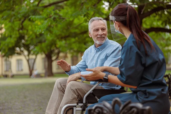 Pria lanjut bicara, memulihkan pasien di kursi roda melakukan percakapan dengan perawatnya dengan pelindung wajah, beristirahat bersama di taman dekat rumah sakit — Stok Foto