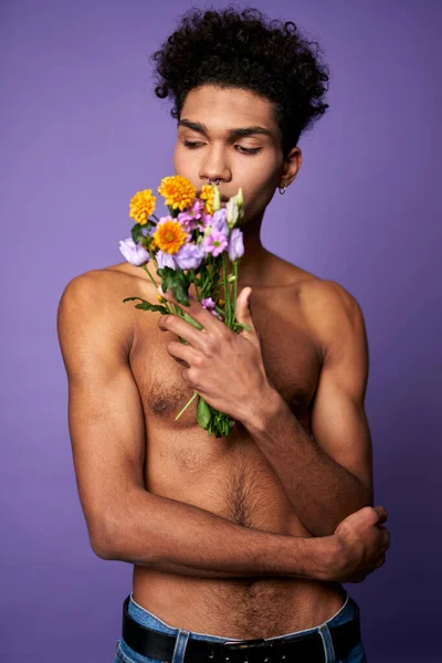 Egy fiatal férfi portréja, lila háttéren pózoló virágokkal. Érzéki transznemű fiú állvány — Stock Fotó