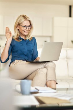 Evde çalışan çekici, olgun bir kadın. İnternetteki video görüşmesi sırasında web kamerasına el sallıyor. Modern apartmandaki bir kanepede otururken laptop kullanıyor.