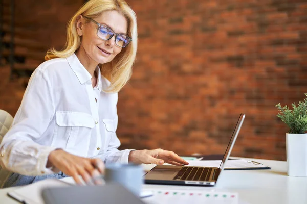 Pengusaha wanita dewasa yang menarik berkacamata menggunakan laptop dan membuat beberapa dokumen di kantor — Stok Foto