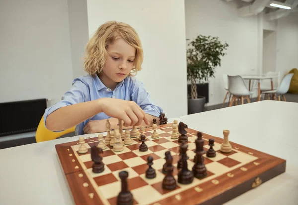 Организованный кавказский мальчик выглядит задумчиво во время игры в шахматы, сидя за столом в школе — стоковое фото