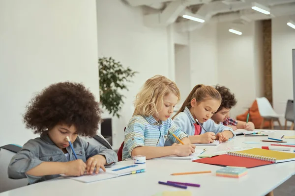 Różnorodne dzieci, małe uczennice robiące notatki, uczące się razem przy stole w szkole podstawowej. — Zdjęcie stockowe