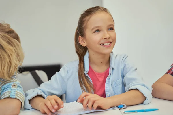 Aranyos kis iskolás lány portréja, amint mosolyog és hallgat, miközben tanul, ül az asztalnál az általános iskolai osztályteremben. — Stock Fotó