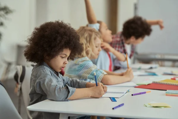 Flitig liten skolpojke skriver i sin anteckningsbok medan han studerar, sitter vid bordet i grundskolans klassrum — Stockfoto