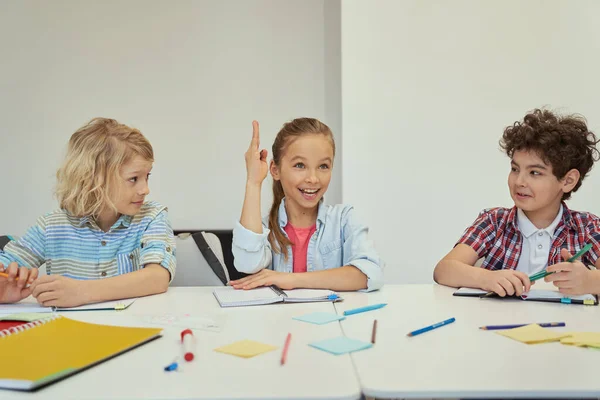 Smart liten skolflicka tittar bort räckte upp handen medan hon studerar, sitter vid bordet i grundskolan klassrum med andra barn — Stockfoto