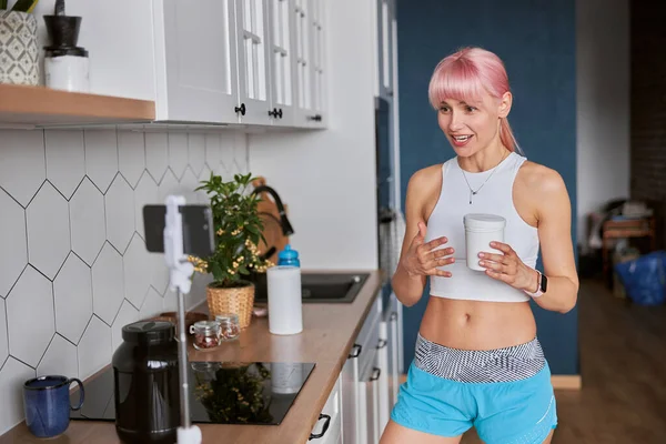 Молодая женщина показывает банку пищевой добавки запись нового видео на кухне — стоковое фото