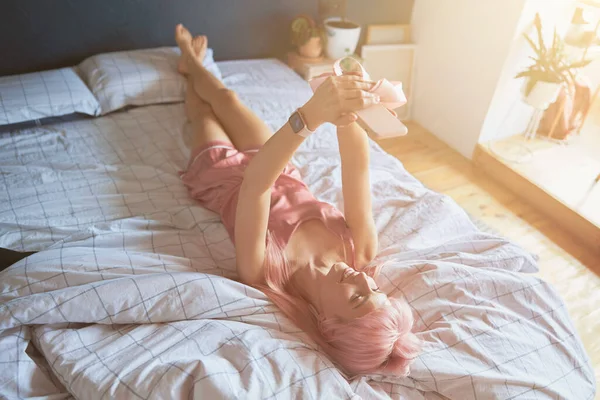 Pretty lady model z kolorowymi różowymi włosami w jedwabnej piżamie robi selfie na wygodnym łóżku — Zdjęcie stockowe