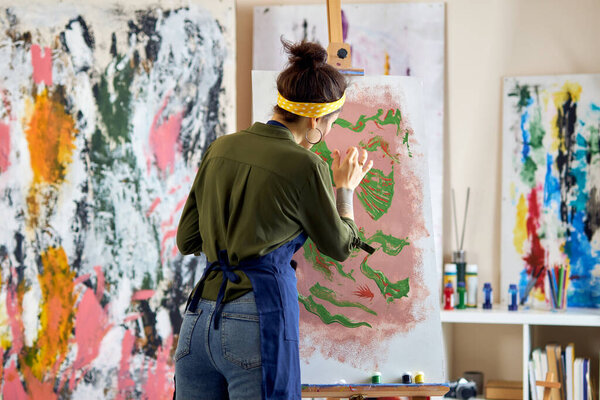 Back view of young female artist in apron painting picture, standing in living room at home