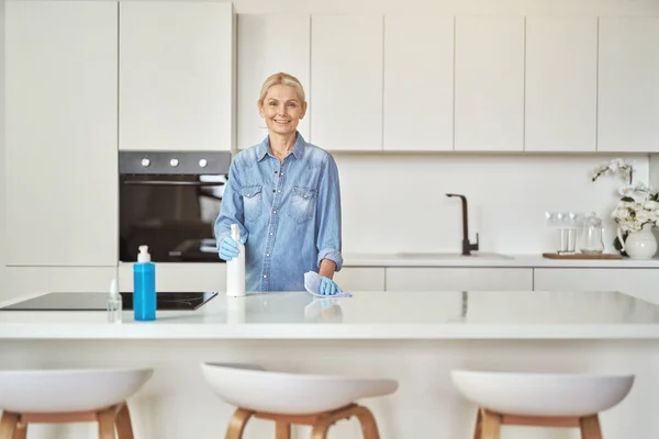 Lastik eldivenlerle gülümseyen olgun ev hanımı gündüz evde deterjanla mutfak yüzeyini temizlerken kameraya bakıyor. — Stok fotoğraf