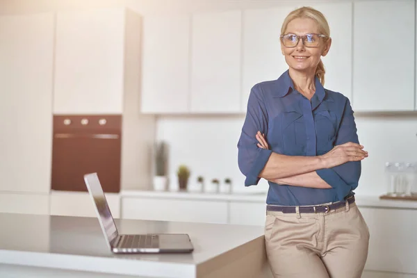Confident mature businesswoman in glasses smiling aside, working at home using laptop, standing with arms crossed in modern kitchen — Stok Foto