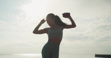 Genç bir kadın okyanusun kenarında neşeyle dans ediyor, hareketlerini ve enerjisini büyüleyici bir zemin üzerinde kucaklıyor..