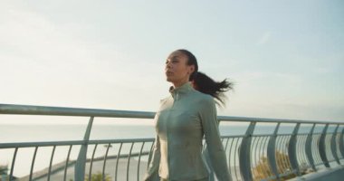 Kendini adamış bir kadın açık havada egzersiz yapıyor, şık giyinmiş ve güçlü hissederken deniz manzarasının tadını çıkarıyor.