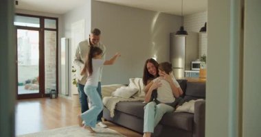 Ebeveynler ve çocuklar oynuyorlar ve açık bir yaşam alanında kanepede kucaklaşıyorlar, enerjik sabah rutini kahkahalarla, zıplamalarla