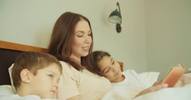 Anne, çocuklarla yatakta kitap okuyor. Hafif lamba örtüleri, nazik anlatımlar. Yatma zamanı, rahatlatıcı atmosfer.