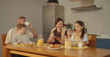 Kafkas ailesi modern mutfakta birlikte kahvaltı yapıyor, iki çocuk ve ebeveyn mısır gevreği, tost ve portakal yerken gülüyor.