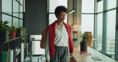 Parlak ofiste duran, masanın yanında uzanan, şehir penceresinden dışarı bakan, omuzlarının üzerinden kırmızı süveter giyen, bitkiler giyen siyah kadın.