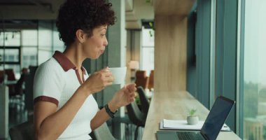 Düzenleme sırasında kahvesini yudumlayan siyah bir kadın, editör bilgisayardaki belgeleri gözden geçiriyor, bitki dolu temiz bir masa, yumuşak gün ışığı, konsantre olmuş.