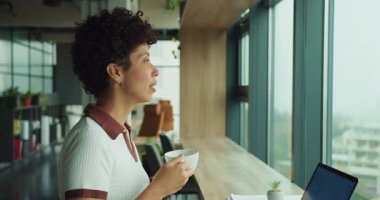 Elinde kahve olan siyah bir kadın. Pazarlama müdürü stratejiyi yansıtıyor. Camdan şehir silueti, yumuşak ofis.