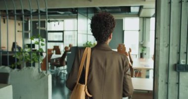 Siyah kadın çantasıyla modern ofise giriyor, açılış planı çalışma alanını, cam duvarları, bitkileri, deri sandalyeleri keşfediyor.,