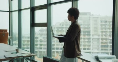 Proje yöneticisi cam ofiste dizüstü bilgisayardaki programı incelerken pencere, kağıt ve çizelgeler masada duruyor.