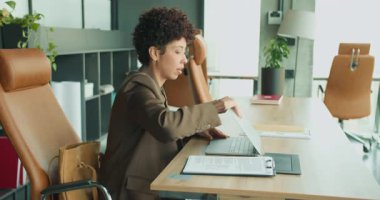 Bilgisayarda, modern ofiste, deri koltukta, bitkilerde, evrakları inceleyen, dosyaları düzenleyen, planlayan siyah bir kadın.