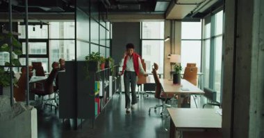 Siyah kreatif direktör çalışma alanında yürüyor, iş arkadaşlarını selamlarken masalar ve bitkiler arasında gidip geliyor, dizüstü bilgisayarları kontrol ediyor.