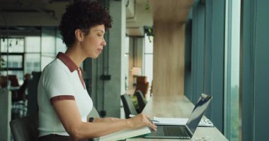 Kadın, pencere önünde dizüstü bilgisayarın yanına notlar yazıyor, deftere fikirler çiziyor, odaklanmış planlama ve proje organizasyonu yapıyor.,