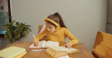 Beyaz kız çalışma masasında detaylı notlar alıyor. Çalışma kitabının kalemine yaslanıyor. Sayfa yoğunlaştırılmış ifade sarı kulaklıklar üzerinde yarışıyor.