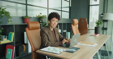 Modern masadaki gülümseyen girişimci dizüstü bilgisayardaki stratejiyi sunuyor, çizelgelere ve belgelere işaret ediyor, deri koltuk, bitki dekoru,