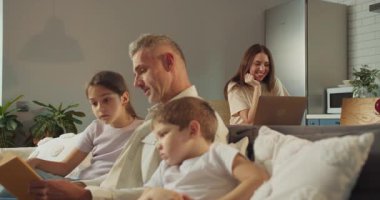 Kafkas ailesinin işi dengelemesi ve babacılık oynaması. Anne dizüstü bilgisayarda yazarken, ilgili çocuklara yüksek sesle kitap okuyor.