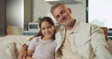 Baba ve kızı birlikte kanepede gülümsüyor. Sakin bir daire ortamında, akşam ışığı pencerelerden süzülüyor.