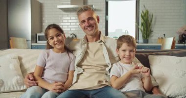 Baba ve çocuklar kanepenin üzerinde gülümsüyor ve rahatlıyor. Modern bir mutfak ortamında baba kızını ve oğlunu sıcak tutuyor.