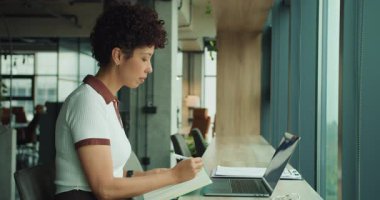 Pencere kenarına notlar yazan kadın, dizüstü bilgisayarı açık fikirler yazan içerik yazarı, yumuşak doğal ışık ve şehir