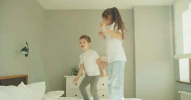 Çocuklar güneş ışığıyla yatakta zıplıyorlar. Yüksek enerjili, neşeli hareketler. Kaygısız, eğlenceli kardeş kıkırdayan şilteler.