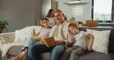 Baba kitap okurken çocuklar kanepede sarılır. Sıcak lamba ışığı, yumuşak battaniye ve hayal gücü ile nazik hikaye anlatma anı.