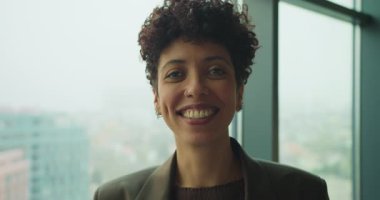 Şehre tepeden bakan pencereye gülümseyen siyah kadın, doğal ışık, özel dikim ceket, doğrudan kamera bakışı, dost canlısı yönetici.